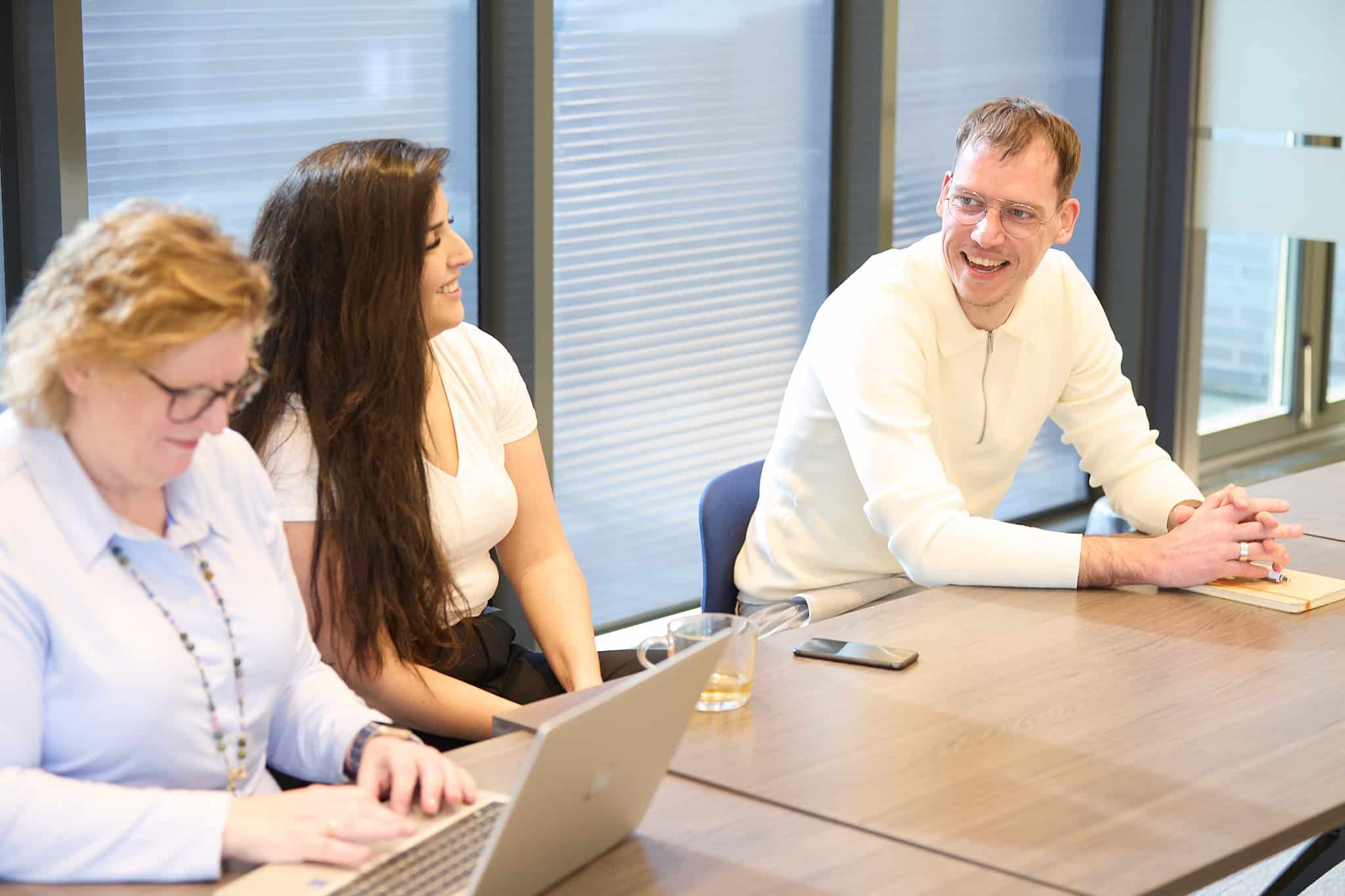 Drie medewerkers overleggen; vrouw typend op laptop, collega’s luisteren en lachen
